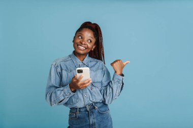 Genç siyahi bir kadın mavi arka planda izole edilmiş bir cep telefonu kullanıyor.