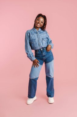 Pembe arka planda izole edilmiş kameraya gülümseyen kot elbiseli genç siyahi kadın.