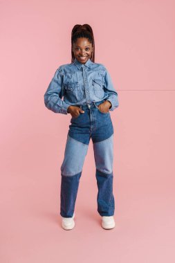 Pembe arka planda izole edilmiş kameraya gülümseyen kot elbiseli genç siyahi kadın.