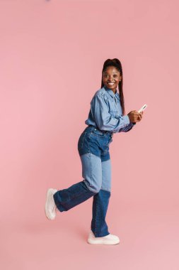 Gülümseyen ve cep telefonu kullanan genç siyah kadın pembe arka planda izole edilmiş.