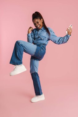 Heyecanlı siyah kadın pembe arka planda izole edilmiş bir şekilde el kol hareketi yapıyor ve cep telefonu kullanıyor.