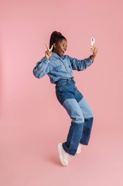Genç siyahi kadın pembe arka planda izole edilmiş cep telefonuyla selfie çekerken işaret veriyor.