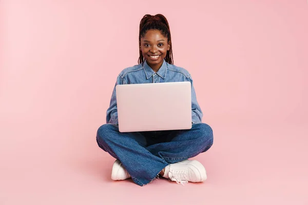 年轻的黑人女人笑着用笔记本电脑 坐在地板上 与粉色背景隔离 — 图库照片