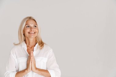 Beyaz arka planda avuçlarını bir arada tutarken gülümseyen olgun sarışın kadın.