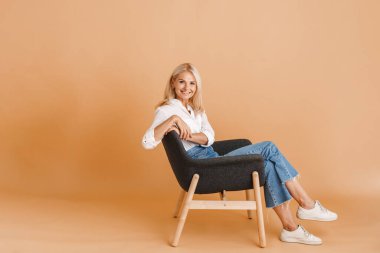 Bej arka planda izole bir şekilde sandalyeye oturmuş kameraya gülümseyen olgun sarışın kadın.