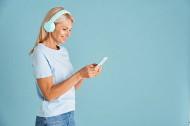 Mavi arka planda izole edilmiş, gülümseyen ve cep telefonu kullanan kulaklıklı olgun sarışın bir kadın.