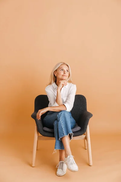 Mature Blonde Woman Looking Aside While Sitting Chair Isolated Beige — Foto de Stock