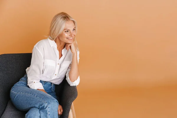 Mature Blonde Woman Smiling Aside While Sitting Chair Isolated Beige — 스톡 사진