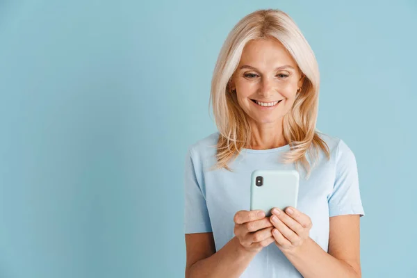 Mature Blonde Woman Wearing Shirt Smiling Using Cellphone Isolated Blue — стоковое фото
