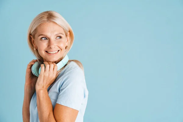 成熟的金发女人 带着耳机 面带微笑 在蓝色的背景下孤零零地望着旁边 — 图库照片