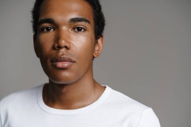 Beyaz tişörtlü genç siyahi adam gri duvarın üzerinde kameraya bakıyor ve poz veriyor.