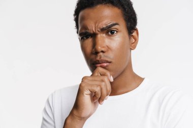 Genç siyah adam kameraya bakıyor ve beyaz duvarın üzerinde çenesini kapalı tutarken kaşlarını çatıyor.