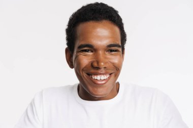 Tişörtlü genç siyah adam gülümsüyor ve beyaz arka planda izole edilmiş kameraya bakıyor.