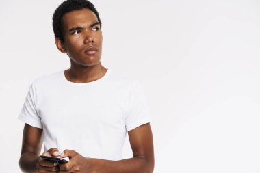 Beyaz duvarda izole edilmiş cep telefonu kullanan genç siyah adam tişörtünü giyiyor.