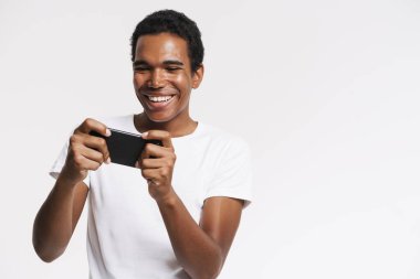 Beyaz duvarda izole edilmiş cep telefonuyla oyun oynarken gülümseyen genç bir zenci.