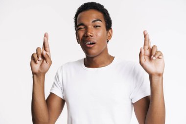 Young black man winking while holding fingers crossed for good luck isolated over white wall