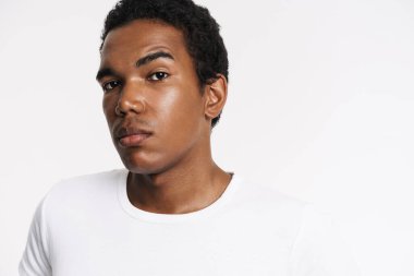Tişörtlü genç siyah adam beyaz arka planda yalnız poz veriyor ve kameraya bakıyor.