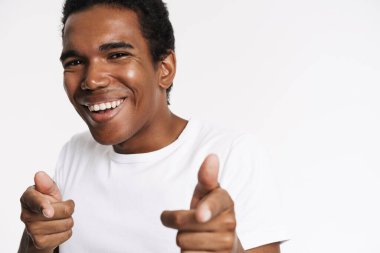 Tişörtlü genç siyah adam gülümsüyor ve beyaz arka planda izole edilmiş kamerayı işaret ediyor.
