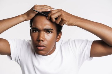 Genç siyahi bir adam beyaz duvarın üzerinde saçlarını tararken kameraya bakıyor.