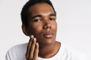Young black man looking at camera while touching his skin isolated over white wall