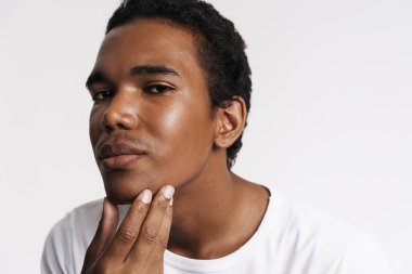 Genç siyah adam beyaz duvarın üzerinden tenine dokunurken kameraya bakıyor.