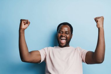 Siyah tüylü adam çığlık atarken kazanan jest yapıyor mavi arka planda izole edilmiş.