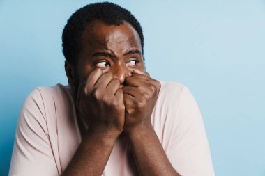 Siyah, korkmuş, tişörtlü, yumruklarını sıkmış, mavi arka planda poz veren bir adam.