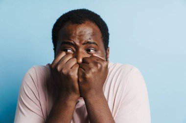 Siyah, korkmuş, tişörtlü, yumruklarını sıkmış, mavi arka planda poz veren bir adam.