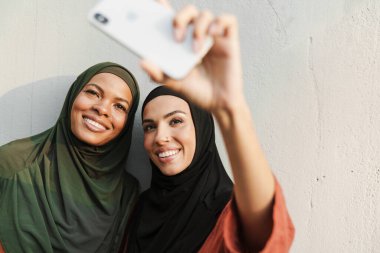 Çok ırklı müslüman kadınlar dışarıda cep telefonuyla selfie çekerken gülümsüyorlar.