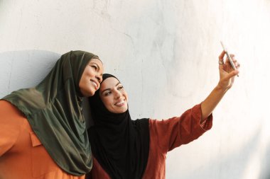 Çok ırklı müslüman kadınlar dışarıda cep telefonuyla selfie çekerken gülümsüyorlar.