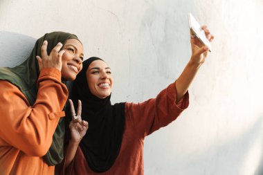 Çok ırklı müslüman kadınlar cep telefonlarında fotoğraf çekerken el kol hareketi yapıyorlar.