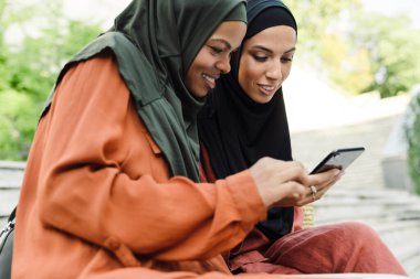 Çok ırklı Müslüman kadınlar açık havada bankta otururken cep telefonu kullanıyorlar.