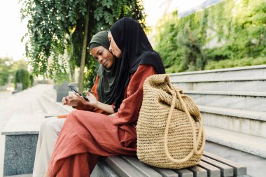 Çok ırklı Müslüman kadınlar açık havada bankta otururken cep telefonu kullanıyorlar.