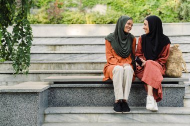 Multiracial muslim women talking and smiling while sitting on bench outdoors