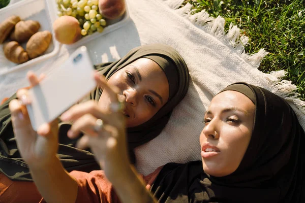 在绿地野餐期间 多种族穆斯林妇女在手机上自拍 — 图库照片