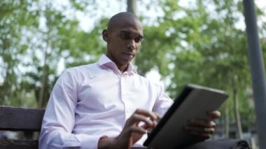 Concentrated African businessman typing by tablet in the park