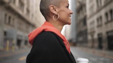 Gülümseyen kel kadın sokakta bir fincan kahveyle yürüyor.