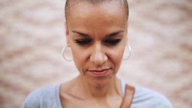 Tişört giyen ve dışarıda kameraya bakan kel bir kadın.