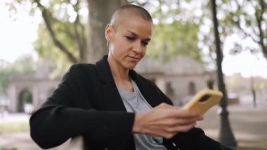 Telefonla yazan ve parktaki bankta oturan kel bir kadın.