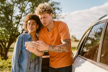 Genç, çok ırklı bir çift yaz gezisinde cep telefonu kullanıyor ve arabada bekliyor.