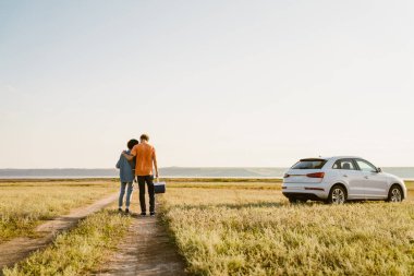 Genç, çok ırklı bir çift araba gezisi sırasında serin bir çantayla sahada dikiliyorlar.