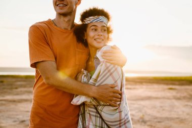 Genç, çok ırklı bir çift dışarıda yan yana dururken sarılıp gülümsüyor.