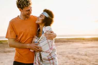 Genç, çok ırklı bir çift dışarıda yan yana dururken sarılıp gülümsüyor.