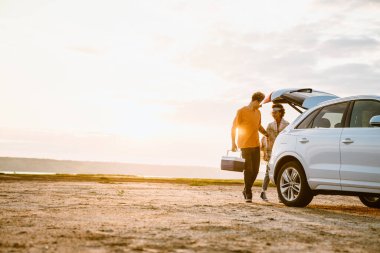 Genç, çok ırklı bir çift araba gezisi sırasında serin bir çantayla sahada dikiliyorlar.