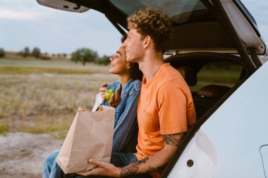 Araba yolculuğu sırasında bagajda oturmuş elma yiyen çok ırklı genç bir çift.