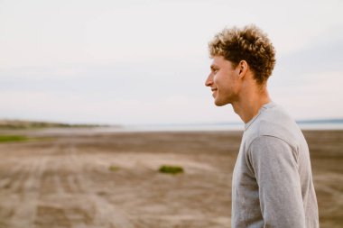 Genç beyaz adam dışarıda dururken gülümsüyor ve yan bakıyor.