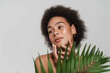 Siyah gömleksiz kadın beyaz arka planda yeşil yapraklı poz verirken yana bakıyor.