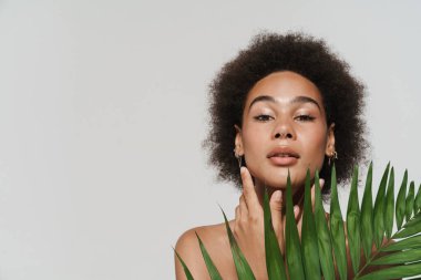 Black shirtless woman looking at camera while posing with green leaf isolated over white background
