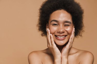 Siyah kıvırcık tişörtsüz kadın gülümsüyor ve bej arka planda izole edilmiş kameraya göz kırpıyor.