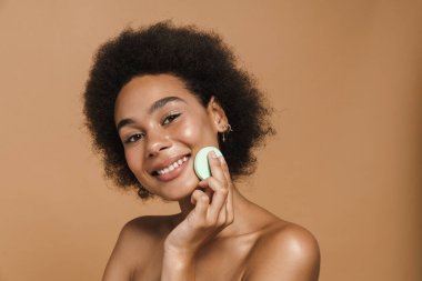 Black curly shirtless woman smiling while using cosmetic sponge isolated over beige background